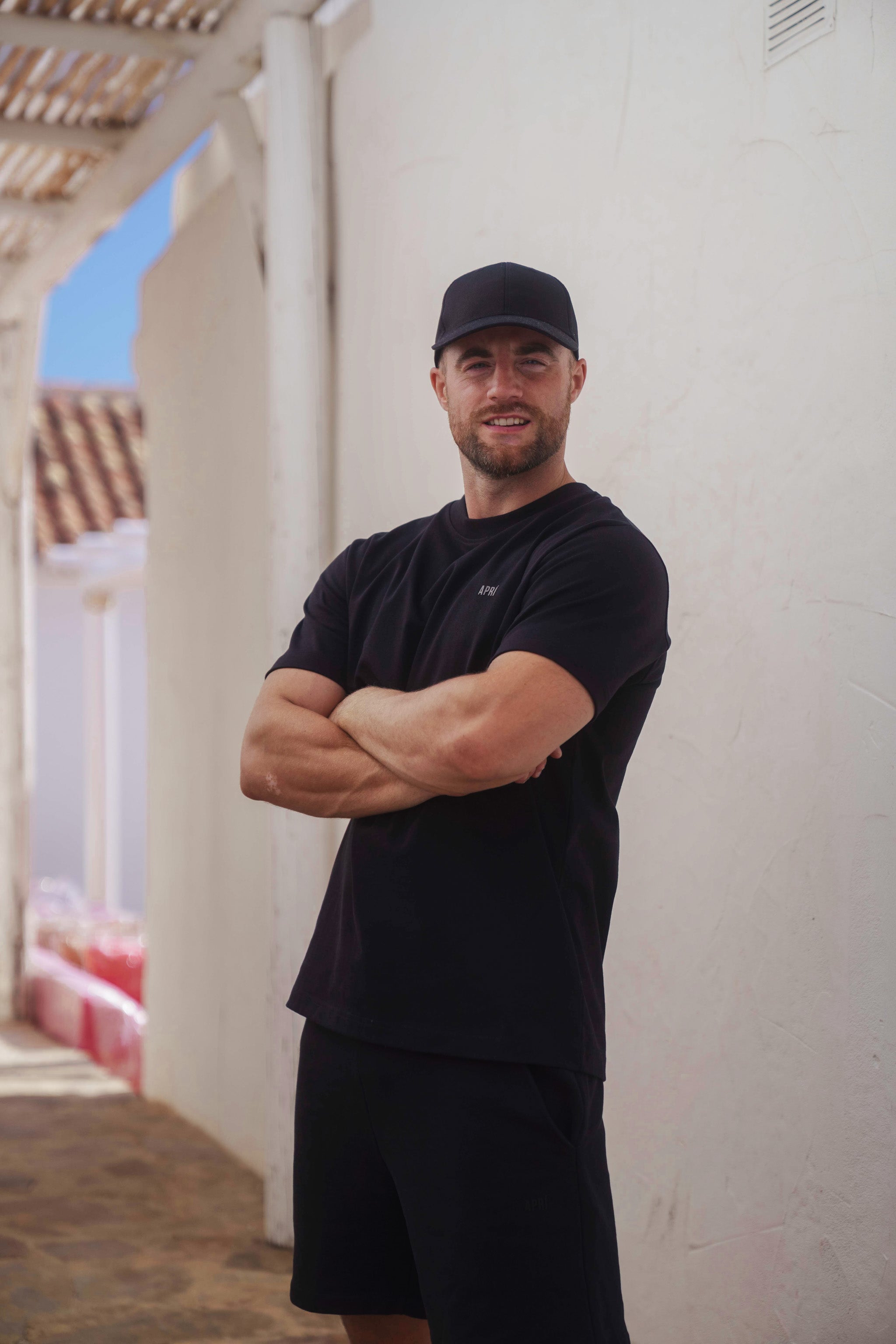 Man wearing a black cap and t-shirt standing against a white wall.